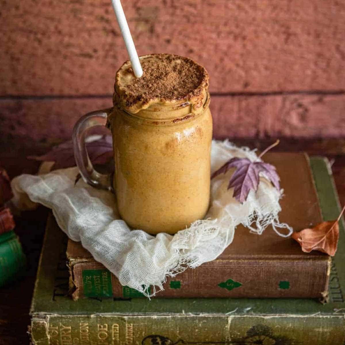 maple pumpkin fall smoothie in a glass mug with a paper straw on top of cheese cloth and a stack of antique books.