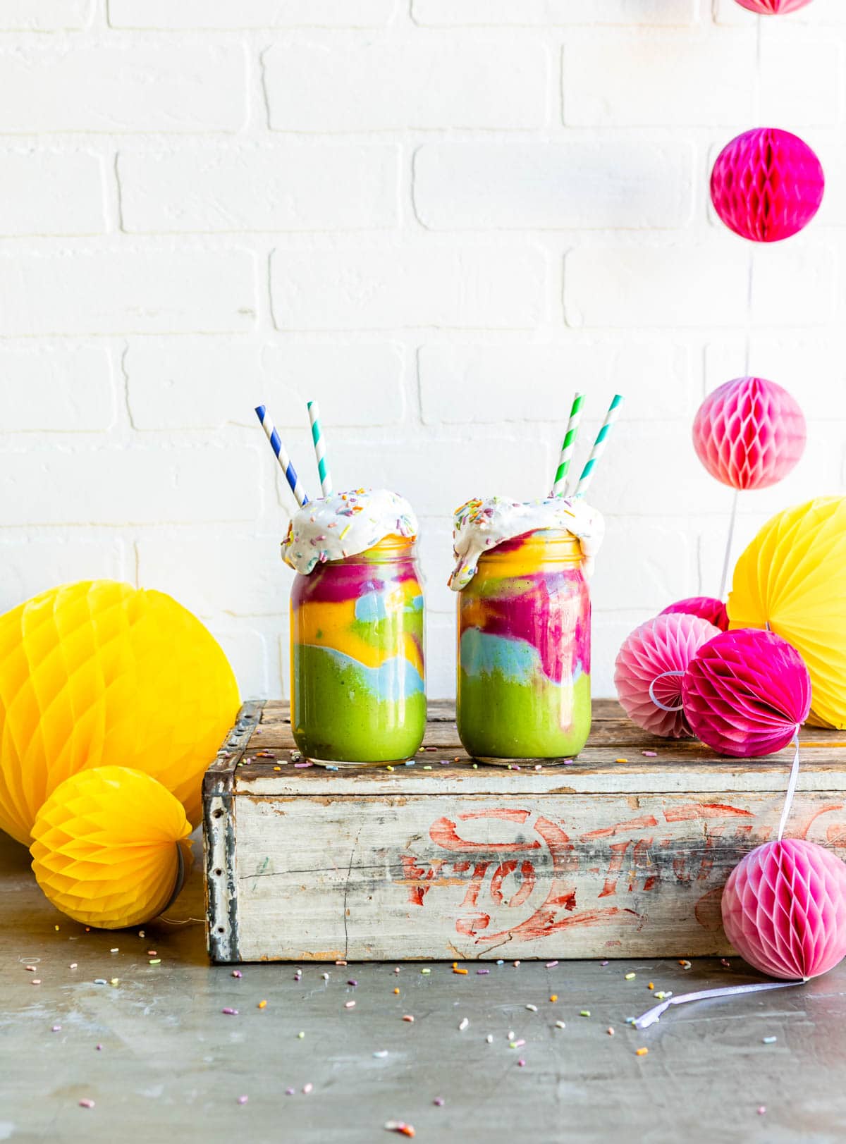 vegan dessert recipe featuring layers of colorful fruit smoothies in jars with coconut whipped cream, sprinkles and paper straws.