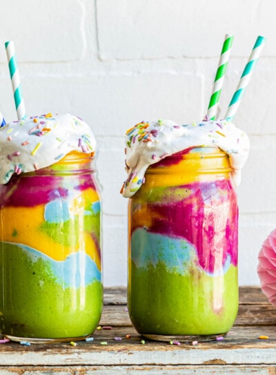 rainbow smoothies in glass jars with coconut whipped cream and paper straws.