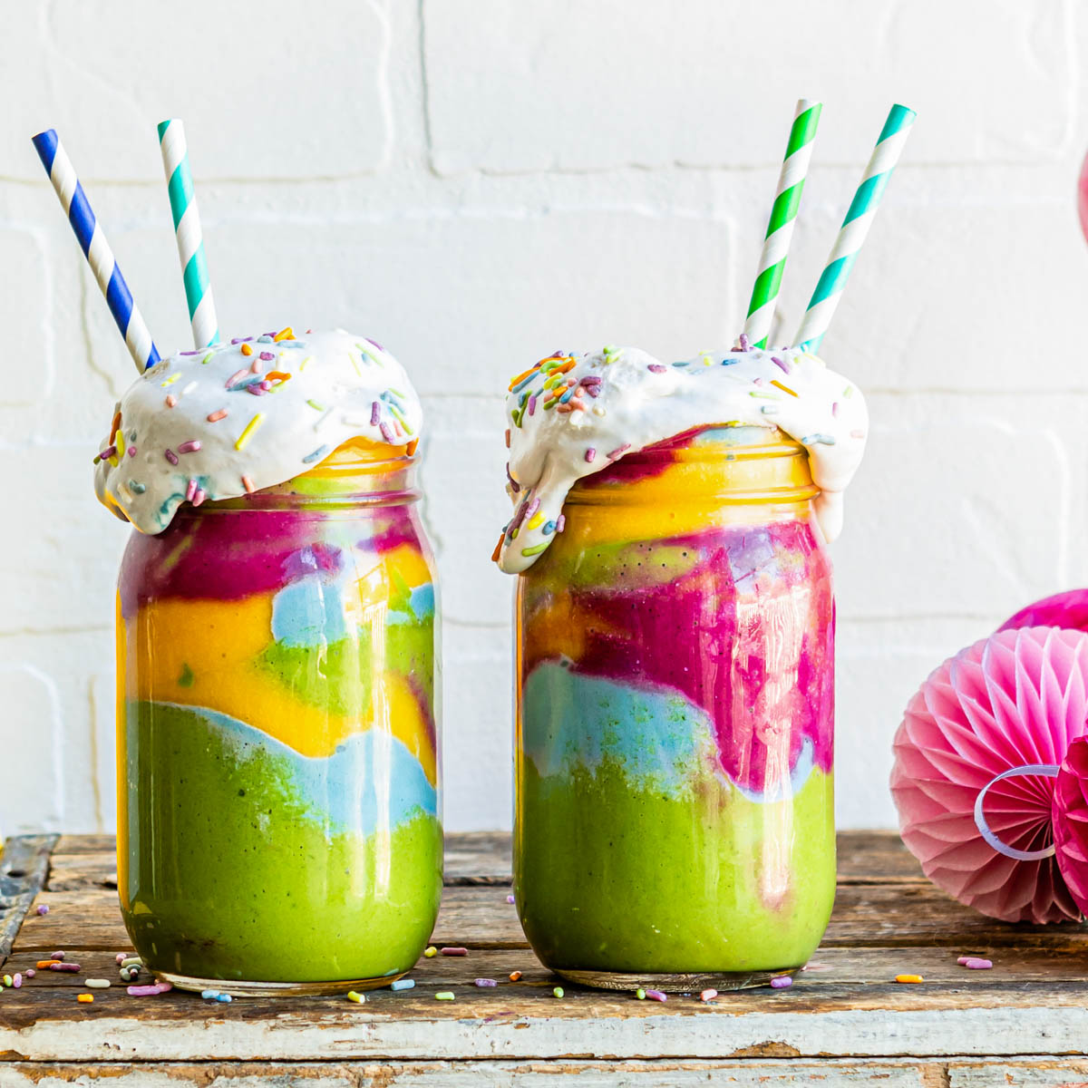 rainbow smoothies in glass jars with coconut whipped cream and paper straws.