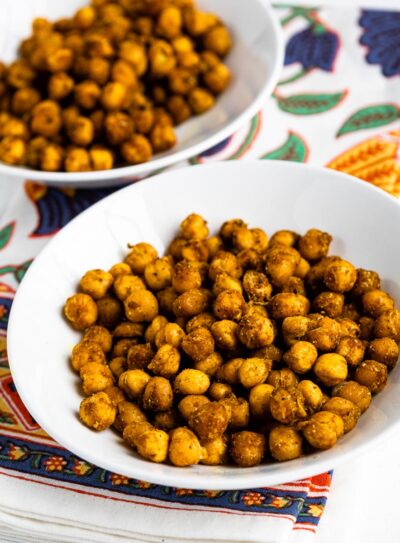 2 white bowls of roasted chickpeas snack on a colorful tea towel.