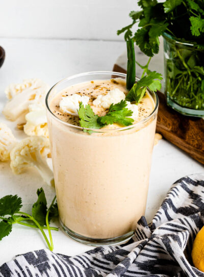 cauliflower smoothie in a large glass topped with seasoned cauliflower florets, green onion and herbs on a white counter top.