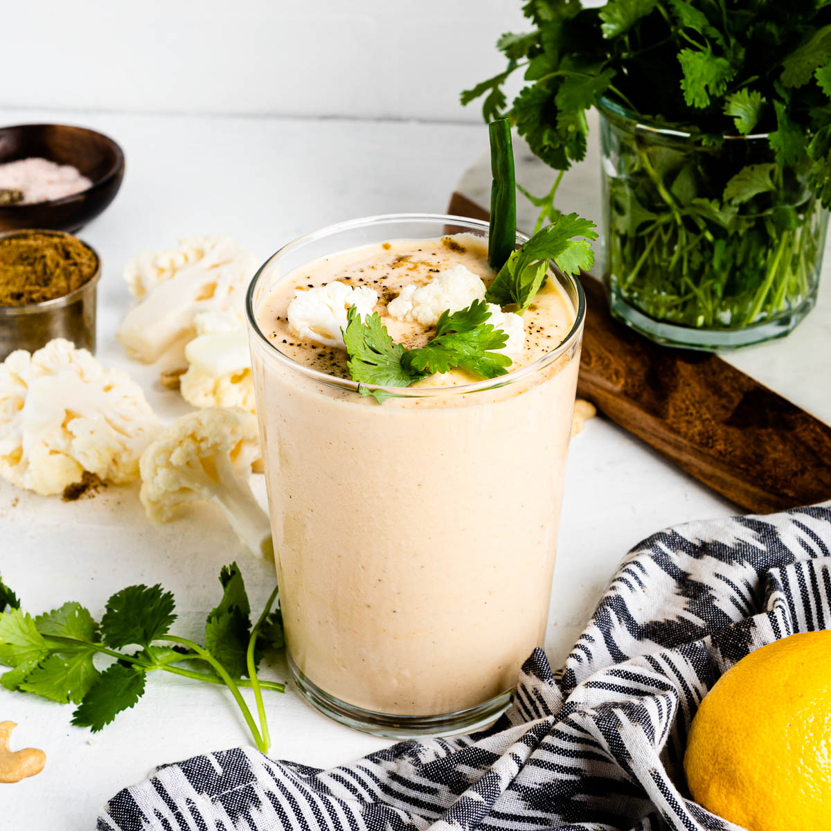 cauliflower smoothie in a large glass topped with seasoned cauliflower florets, green onion and herbs on a white counter top.