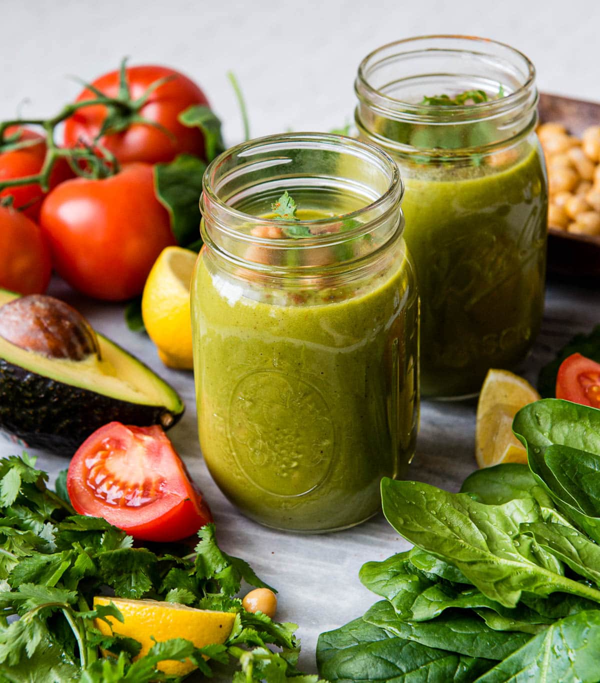 2 glass jars full of green smoothie surrounded by fresh vegetables like tomatoes, avocado and spinach.