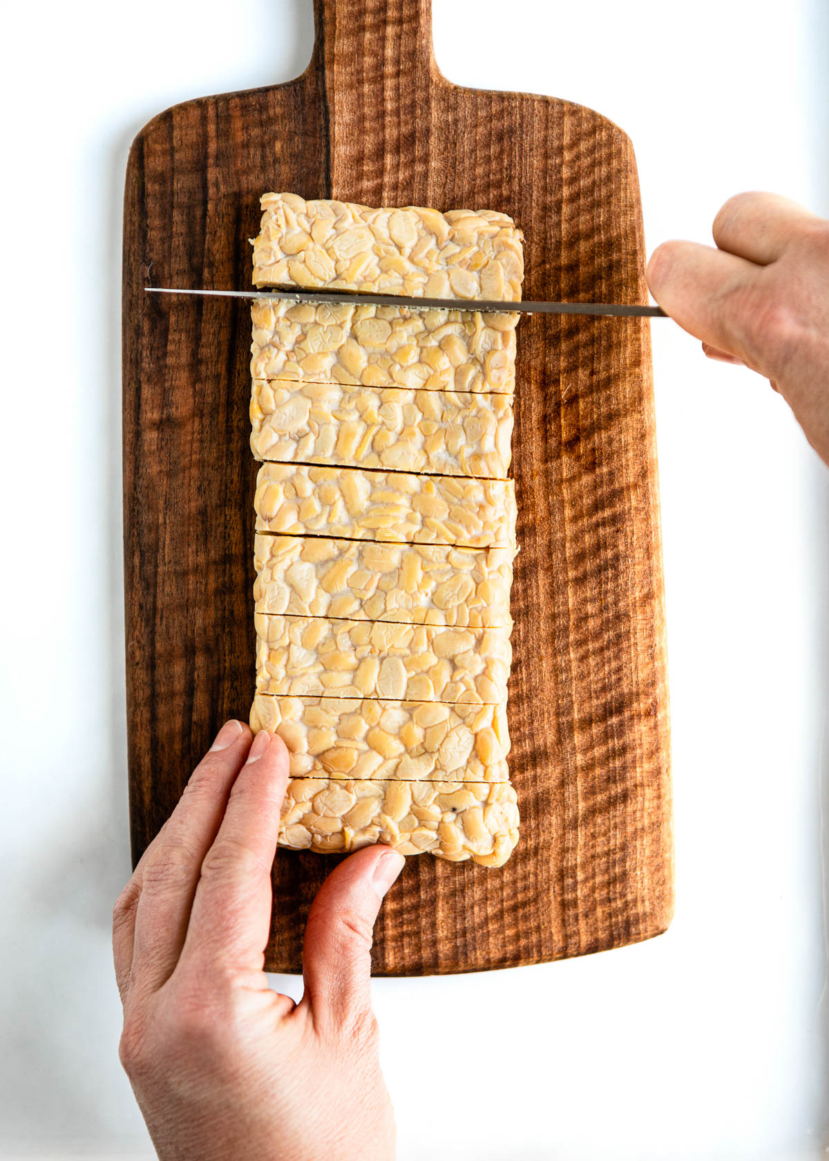 two hands using a chef's knife to make even rectangles of tempeh on a wooden cutting board.