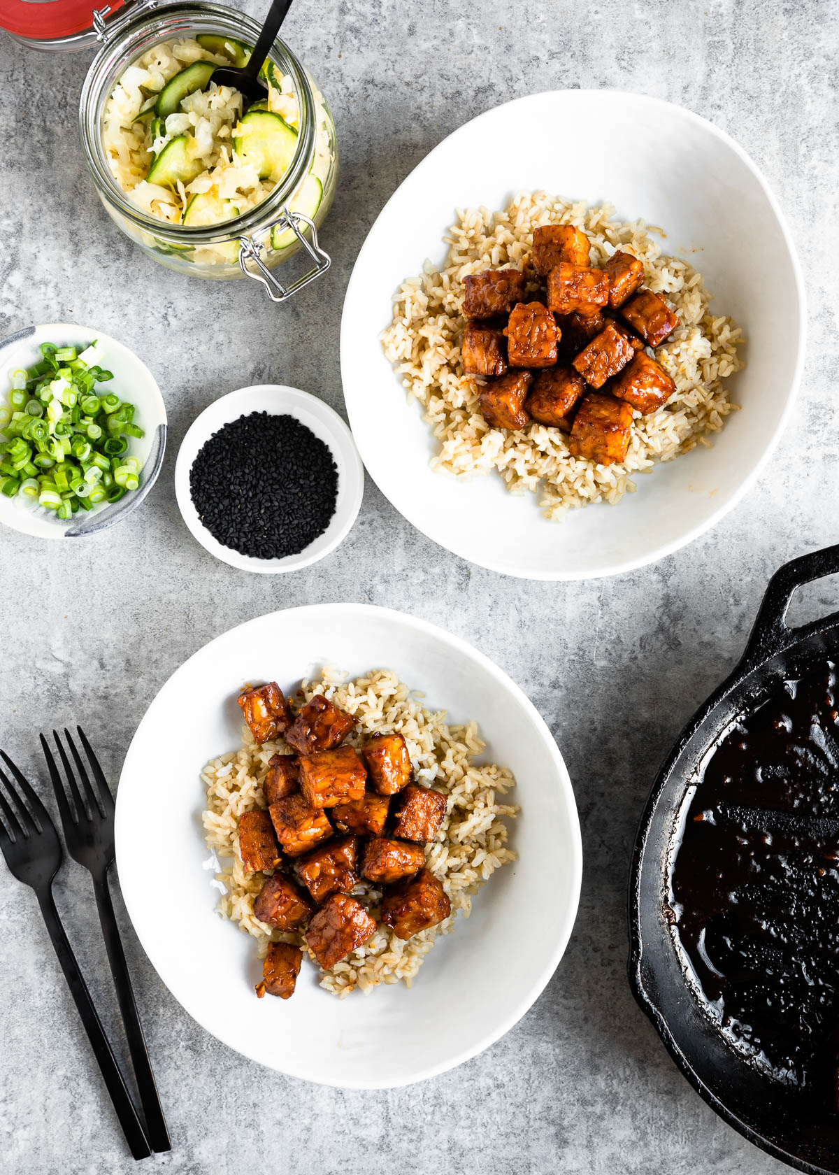 two white bowls filled with brown rice and pieces of cooked tempeh next to containers of toppings including green onion, sesame seeds and pickled vegetables.