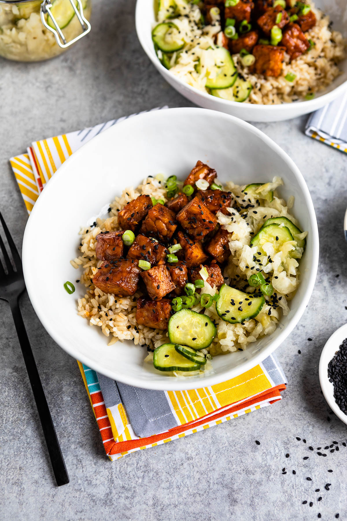 white bowl with brown rice, marinated tempeh and pickled cucumbers all topped with sesame seeds.