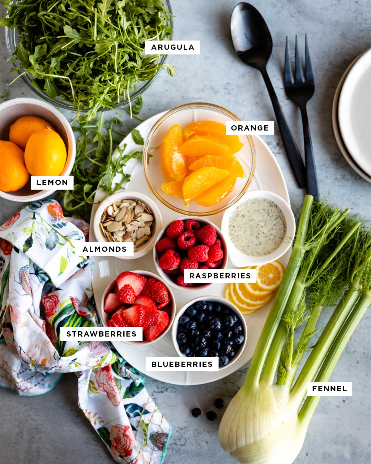 ingredients for strawberry lemon arugula salad in black writing and white text boxes including arugula, lemon, orange, almonds, raspberries, strawberries, blueberries and fennel.