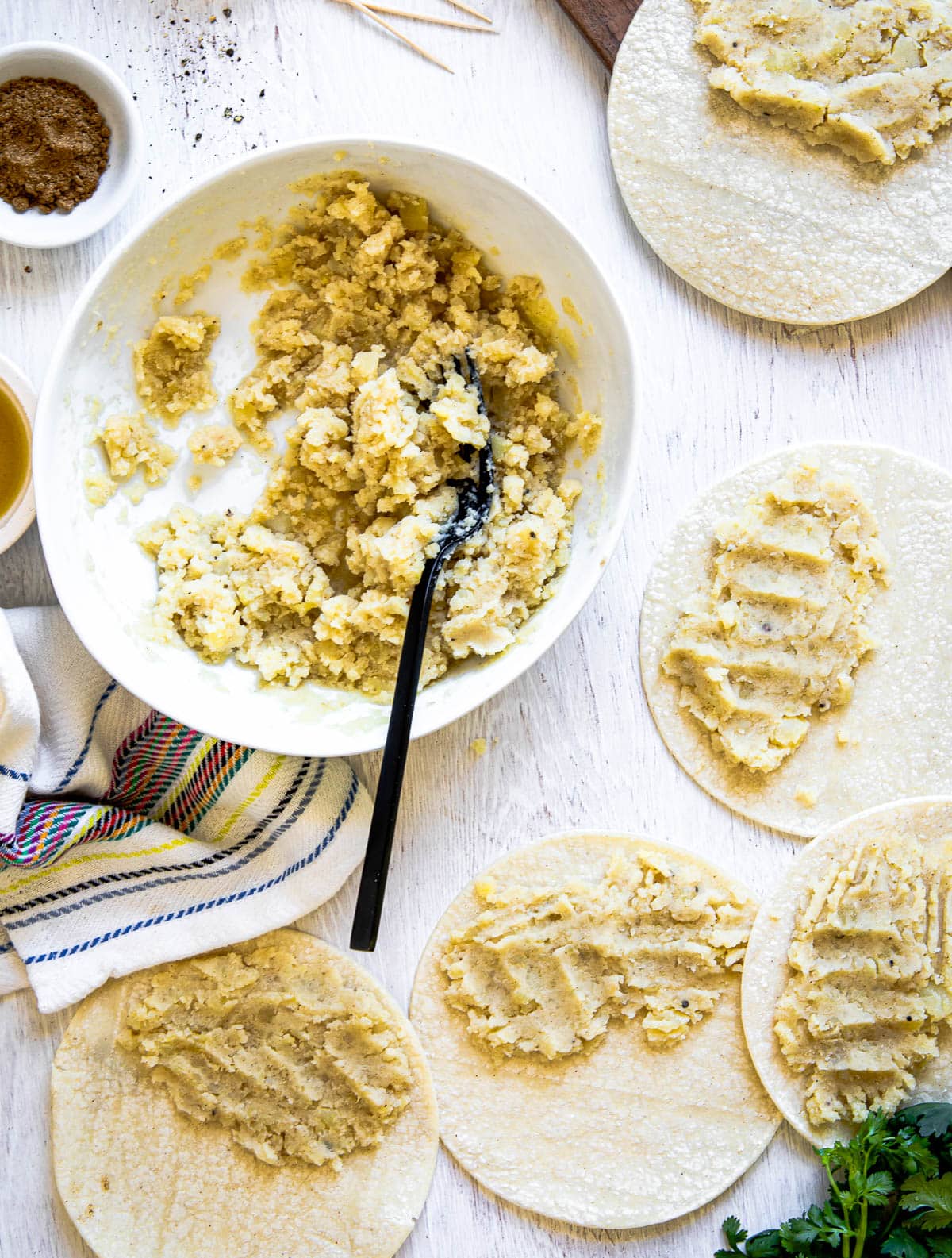 Preparation of tacos de papa recipe with golden mashed potatoes being added to corn tortillas on a white surface.