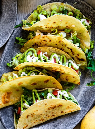 Plate of tacos de papa with crispy potato-filled corn tortillas, topped with fresh vegetables and creamy sauce on an oval, gray plate.