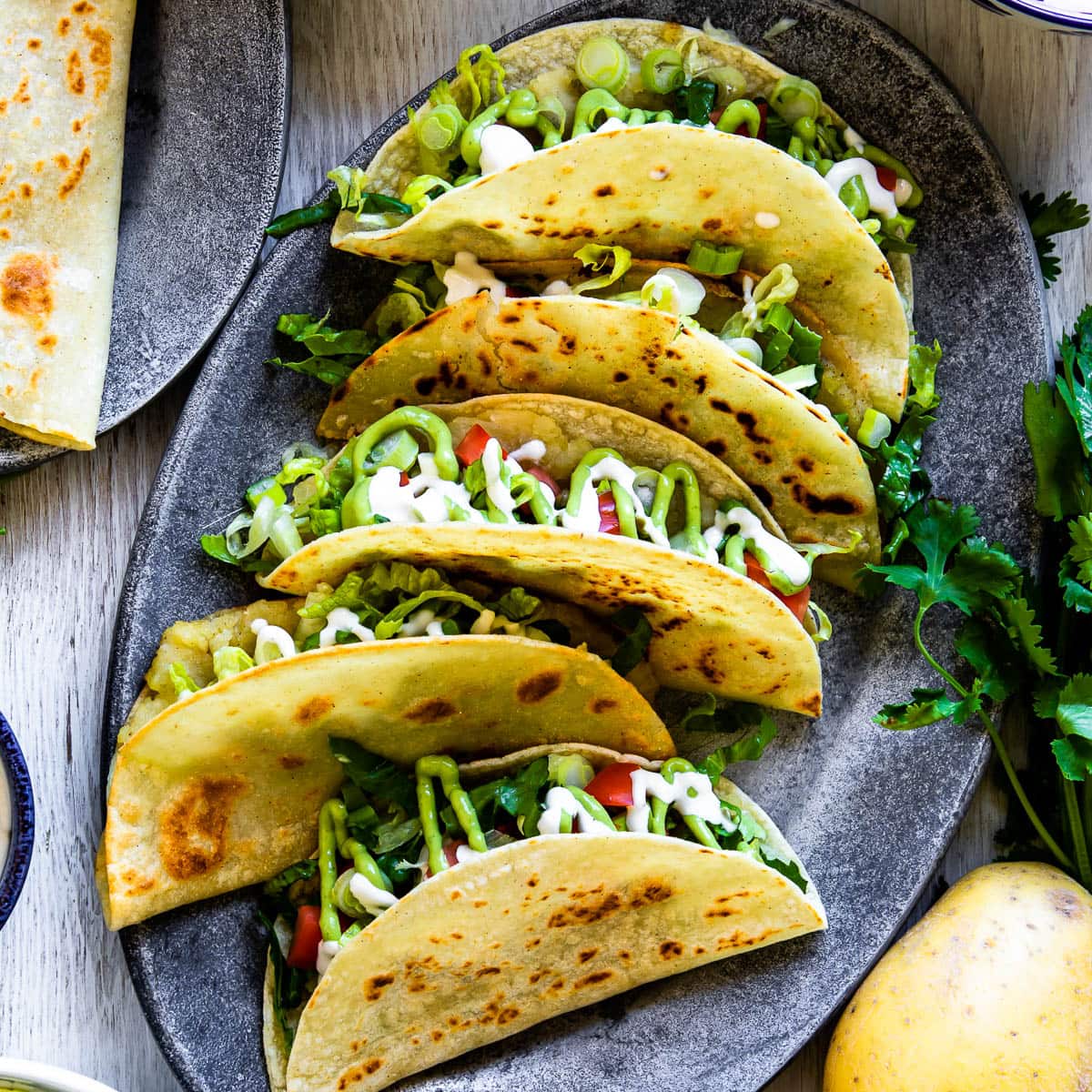 Plate of tacos de papa with crispy potato-filled corn tortillas, topped with fresh vegetables and creamy sauce on an oval, gray plate.