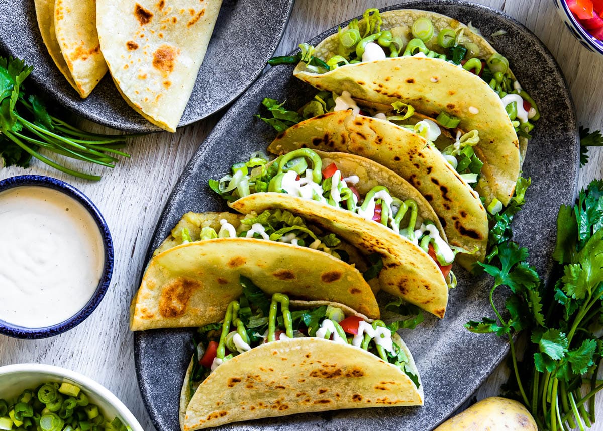 Gray oval plate of tacos de papa topped with avocado crema and cashew cream, showcasing a delicious Mexican tacos recipe.