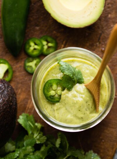 Vegan avocado crema in a glass jar topped with cilantro, sliced jalapeño and a wooden spoon.