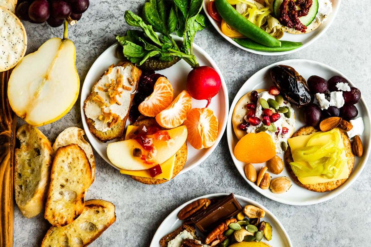 small white plates with appetizers of crackers, dried and fresh fruits, vegan cheeses, nuts, seeds and crusty bread.