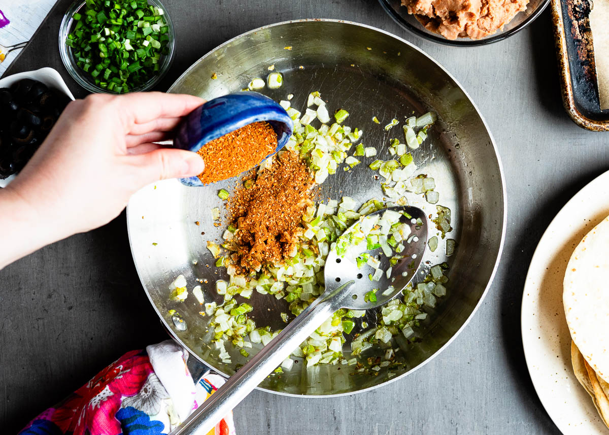 pouring taco seasoning from a small blue bowl into a stainless steel skillet of sautéd vegetables.