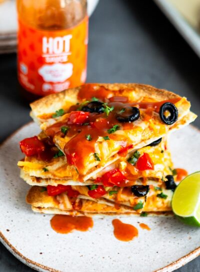 stacked slices of Vegan Mexican Pizza on stone plate next to a bottle of Taco Bell Hot Sauce.