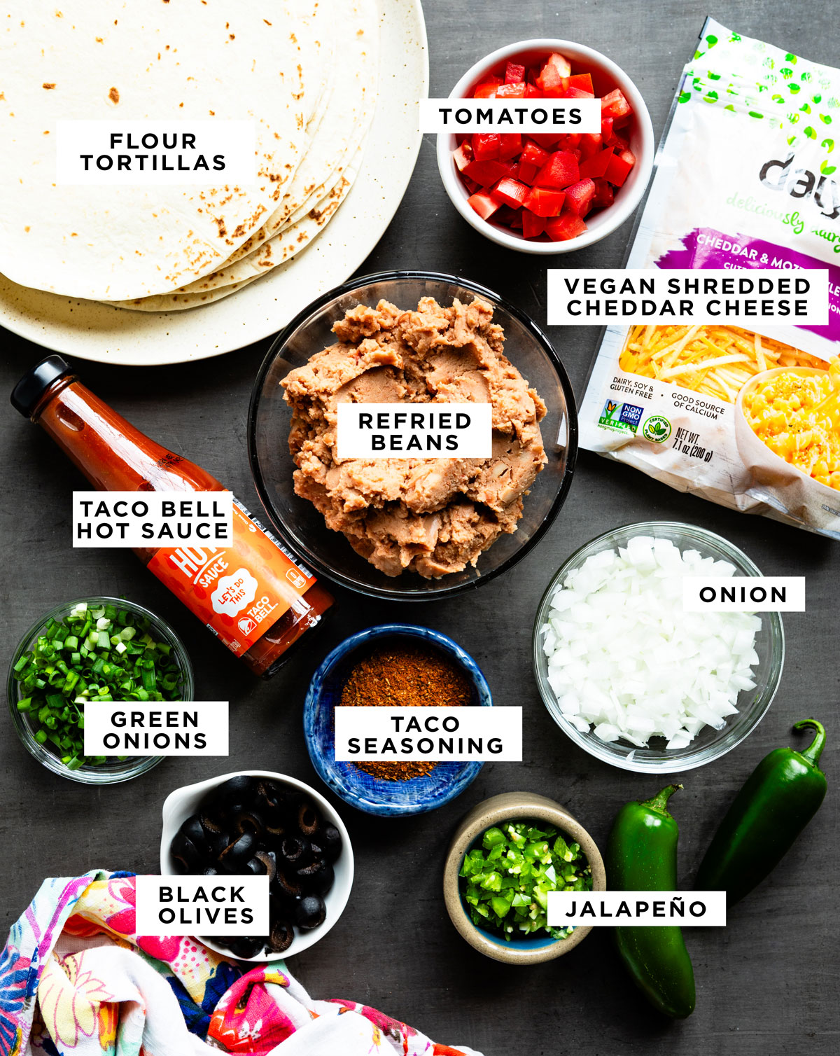 labeled ingredients for vegan Mexican pizza including flour tortillas, tomatoes, vegan shredded cheddar cheese, refried beans, Taco Bell hot sauce, onion, taco seasoning, green onions, black olives and jalapeño.