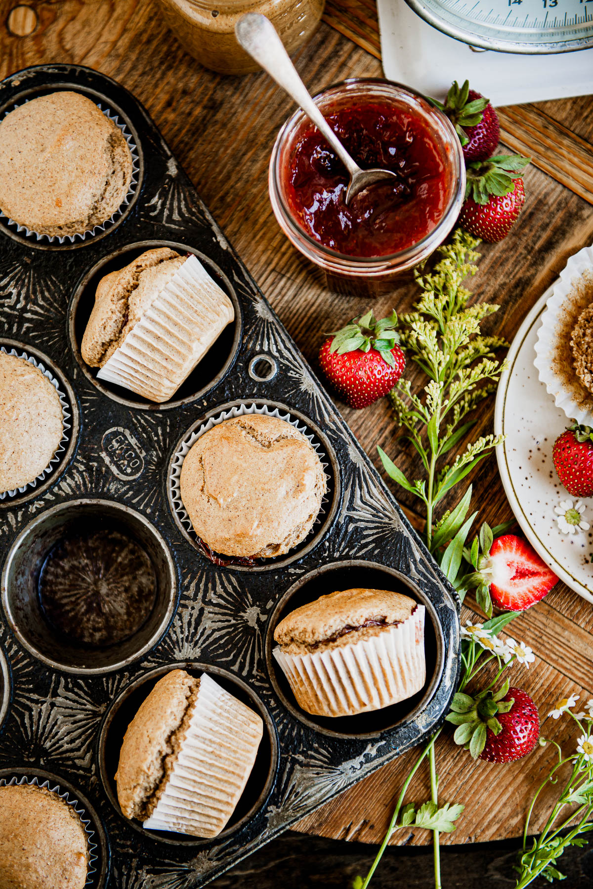 antique black muffin tin with baked vegan muffins in white wrappers, next to strawberry jam.