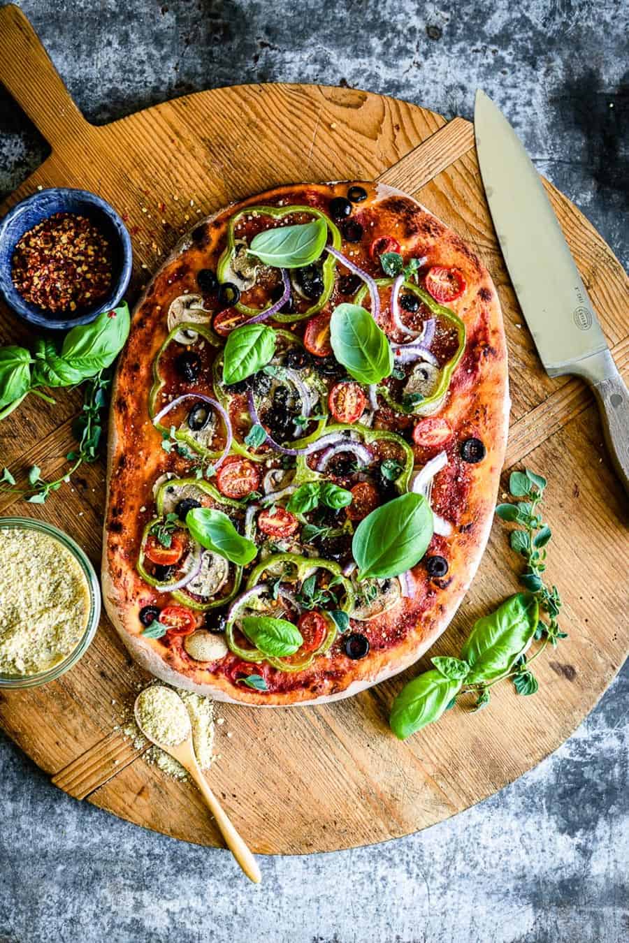 vegan pizza with fresh toppings on a wooden table next to a knife and crushed red pepper.