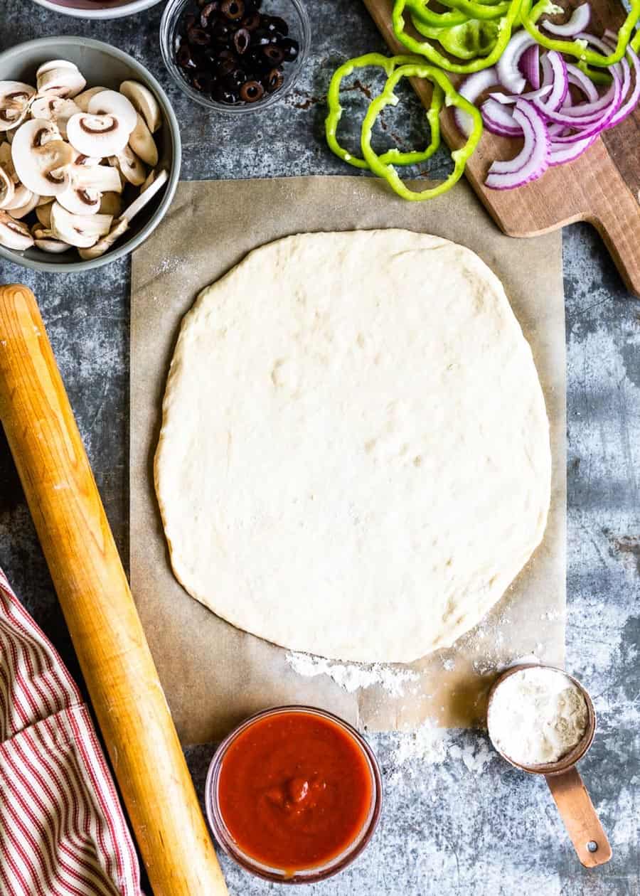 sliced vegetables above a parchment paper with a rolled out pizza dough and a bowl of pizza sauce.