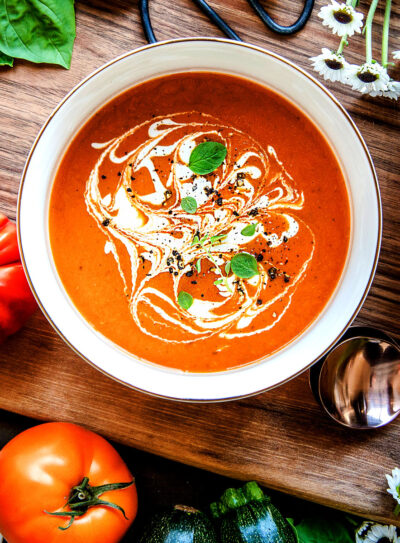 Two bowls of vegan tomato soup topped with vegan cream and basil leaves on a wooden table next to two spoons.