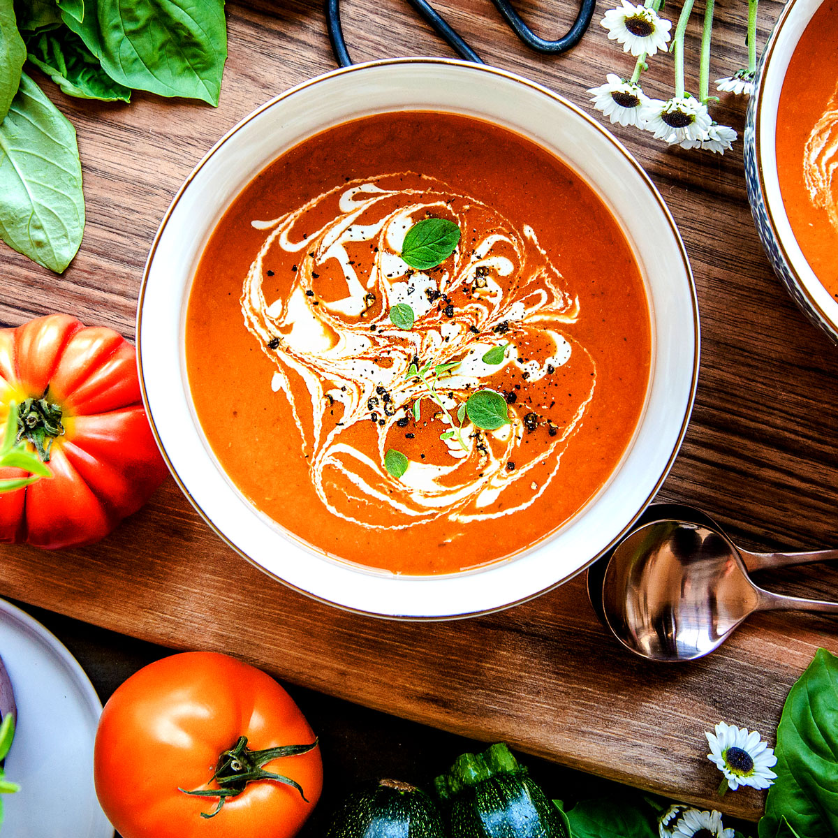 Two bowls of vegan tomato soup topped with vegan cream and basil leaves on a wooden table next to two spoons.