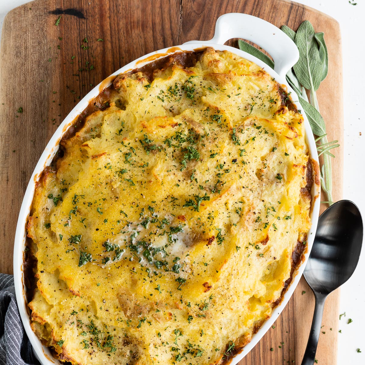 white casserole dish with vegetarian shepherd's pie on a wooden cutting board next to a black serving spoon.