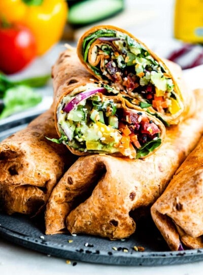 Sliced vegetable wrap placed on top of 3 full wraps, in sun-dried tomato tortilla on a plate. Veggie wrap ingredients are blurred in the background.