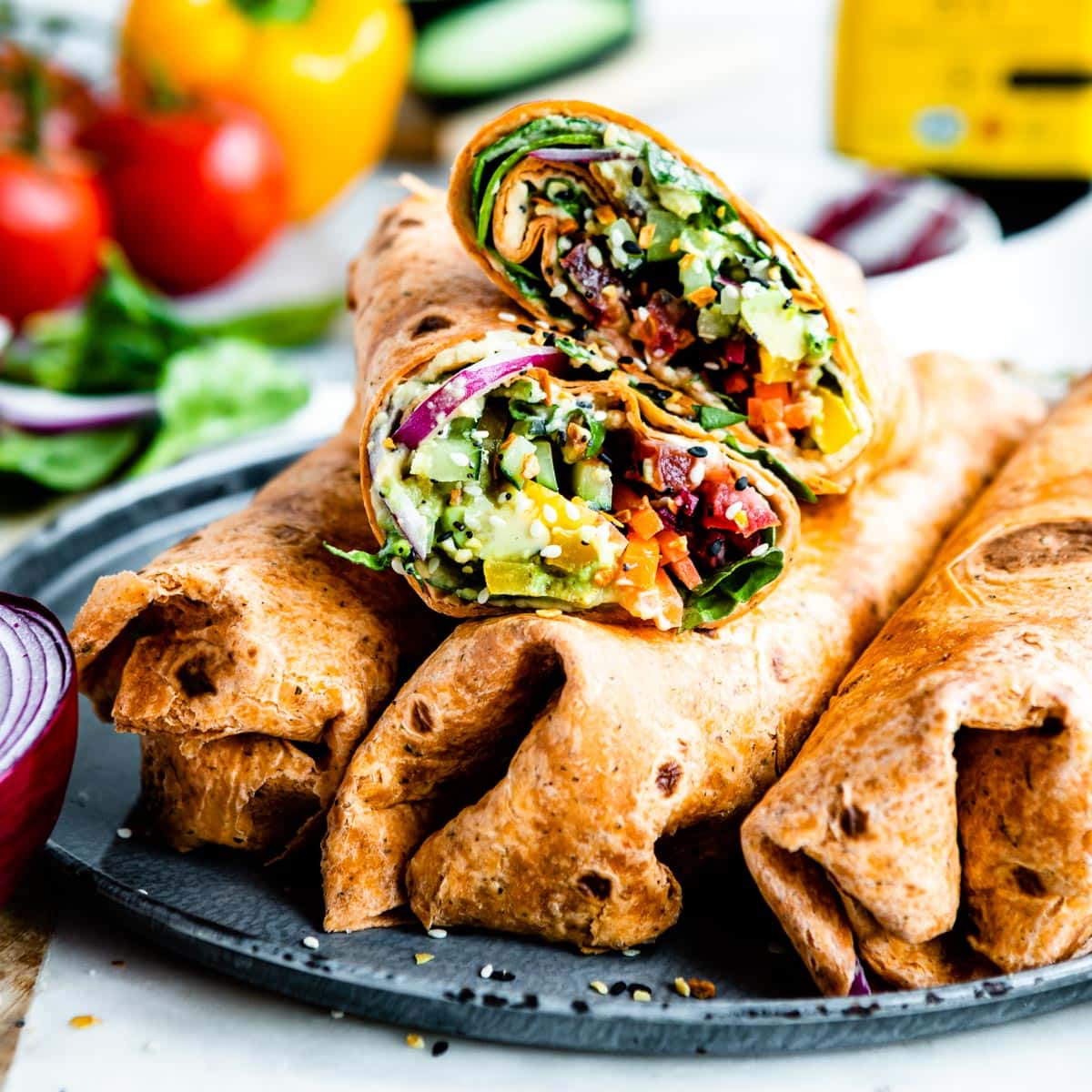 Sliced vegetable wrap placed on top of 3 full wraps, in sun-dried tomato tortilla on a plate. Veggie wrap ingredients are blurred in the background.