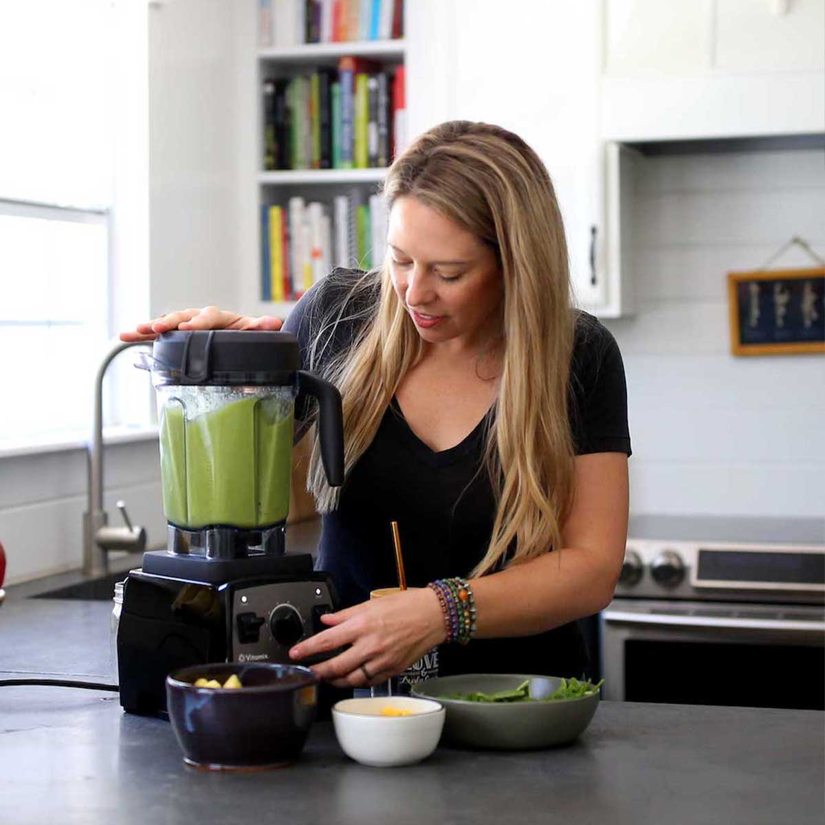 white woman using a vitamix blender to make a green smoothie in a kitchen.