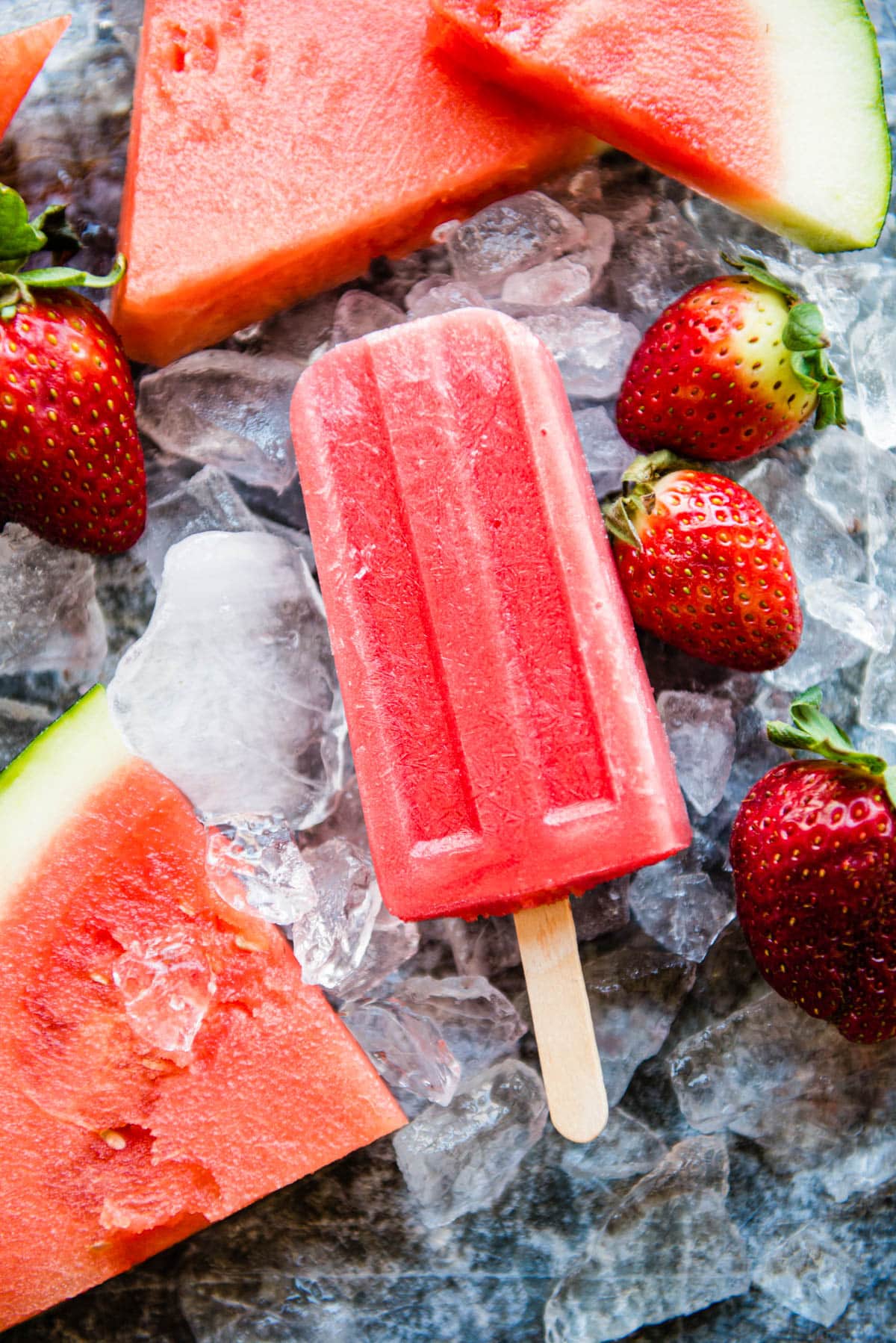 strawberry watermelon popsicles on a wooden stick sitting on ice.