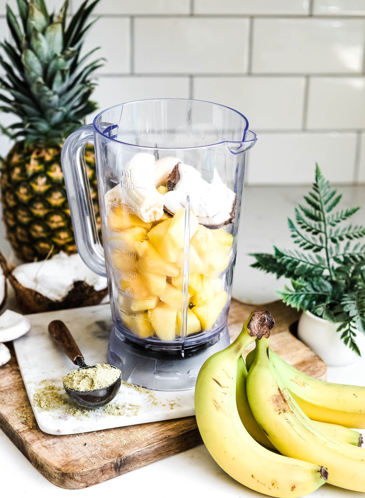 blender container with ingredients for a smoothie on a counter next to a scoop of protein powder and a bunch of bananas.